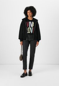 Čierna mikina na zips cez grafické čierne tričko s farebným textom "VIVA LOVE", skombinovaná s tmavými džínsami a čiernymi mokasínmi. Malá hnedá crossbody taška.