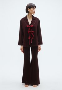 Bordeauxfarvet loungestel med en skjortetop med krave og tre røde båndsløjfer samt vide bukser, lavet af et blødt, tekstureret stof.