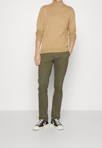Maglione beige a collo alto, pantaloni verde oliva e sneaker nere con accenti bianchi. Design semplice, silhouette aderente, branding minimale.