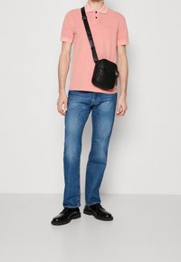Chemise polo rose, jeans bleus et chaussures habillées noires. Un petit sac en bandoulière noir avec une fermeture éclair est porté sur l'épaule.