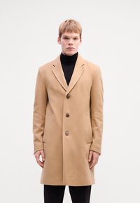 Light beige wool coat with a tailored fit, notched lapels, and three buttons. Features side pockets and a smooth texture.