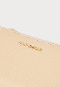 Bežs teksturēts ādas maks ar zeltītu "Coccinelle" logo. Tam ir gluda rāvējslēdzēja mala gar augšējo mali. Kompakts un minimālistisks dizains.