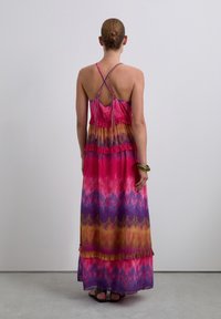 Vrouw met een bun-kapsel, gekleed in een mouwloze, kleurrijke tie-dye maxi-jurk met gekruiste banden en sandaal, staand tegen een effen achtergrond.