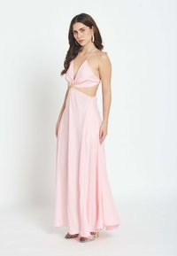 Mujer con cabello largo y oscuro que lleva un vestido maxi color rosa claro con escote halter y aberturas, y sandalias de tacón multicolores, posando contra un fondo blanco.