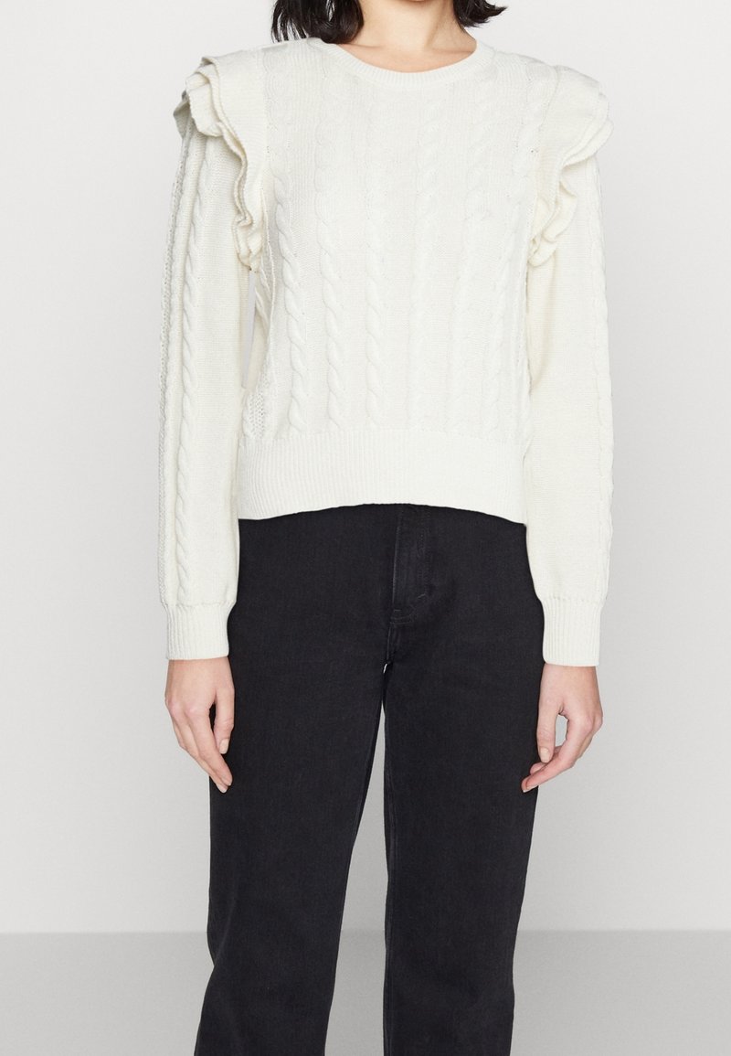 Pull blanc en maille torsadée avec des détails à volants aux épaules, porté avec un pantalon noir taille haute, montré du cou aux hanches.