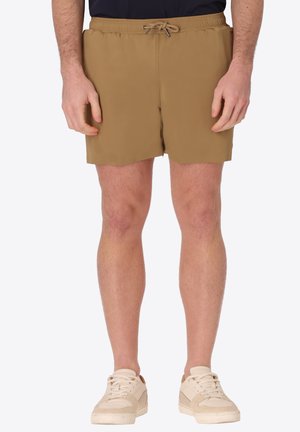 Uomo che indossa pantaloncini beige con coulisse e sneakers bianco sporco in piedi davanti a uno sfondo semplice.