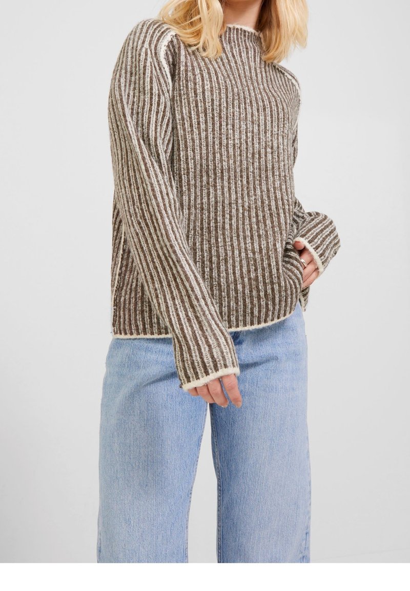 Personne portant un pull en tricot côtelé marron et blanc à manches longues et un jean large bleu clair, sur un fond uni.