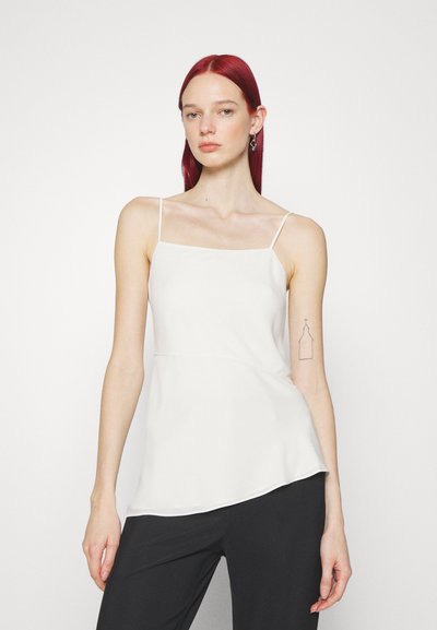 Camisole blanche avec des bretelles fines, texture lisse et ourlet asymétrique, associée à un pantalon noir. Le modèle a les cheveux roux et un tatouage visible.