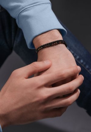 Braccialetto nero intrecciato con perline rotonde su un polso, indossato con una manica lunga azzurro chiaro. Mani che si toccano su uno sfondo di tessuto denim.