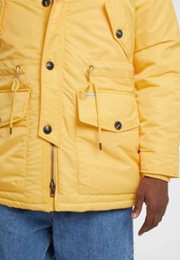 Geel gevoerd jack met rits, drukknoopsluiting, twee voorzakken en trekkoorddetails. Stof heeft een gladde textuur en is gecombineerd met denim.