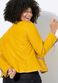 Chaqueta de cuero amarilla con un diseño ajustado, costuras visibles y puños con cremallera. La textura es suave y brillante.
