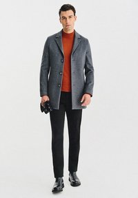 Szary wełniany płaszcz z trzema guzikami i klapami z wycięciem, noszony na pomarańczowym golfie, zestawiony z czarnymi spodniami i czarnymi butami.