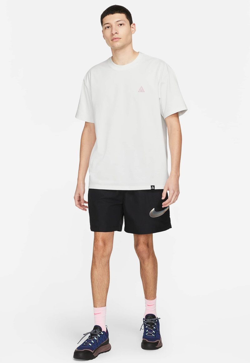 Jeune homme debout, portant un t-shirt blanc à manches courtes, un short noir avec un logo Nike, des chaussettes roses et des baskets bleues.