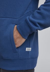 Sudadera azul con dobladillo y puños acanalados, que cuenta con un bolsillo lateral y una pequeña etiqueta de tela en la parte inferior. Textura suave.