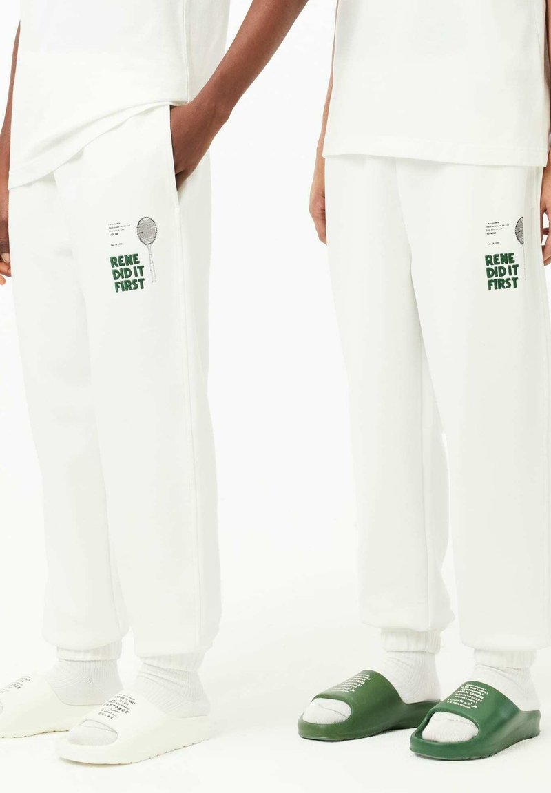 Pantalons de survêtement blancs avec des poignets côtelés, comportant le texte vert "RENE L'A FAIT EN PREMIER" et un graphique. Portés avec des sandales à enfiler blanches et vertes.