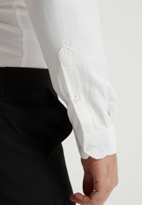Chemise blanche en tissu lisse, dotée de poignets boutonnés et d'une coupe ajustée, associée à un pantalon noir.