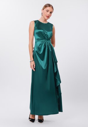 Robe maxi en satin vert émeraude avec un design sans manches, des volants latéraux, une taille froncée et une texture lisse et brillante. Chaussures noires visibles.