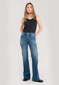 Jean évasé taille haute en denim bleu mi-ton avec des détails décolorés. Associé à un haut sans manches noir et des chaussures noires.