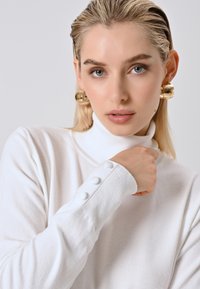 Frau mit nach hinten gekämmtem blonden Haar, die große goldene Ohringe und einen weißen Rollkragenpullover mit Knöpfen an den Ärmeln trägt.