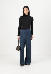 Svart langermet topp og plisserte teal vide bukser. Modellen holder en liten brun håndveske, iført dyreprint flats. Hvit bakgrunn.