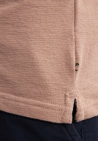 Světle růžová texturovaná košile s jemným vzorem, obsahující boční rozparky a malou etiketu na lemu.
