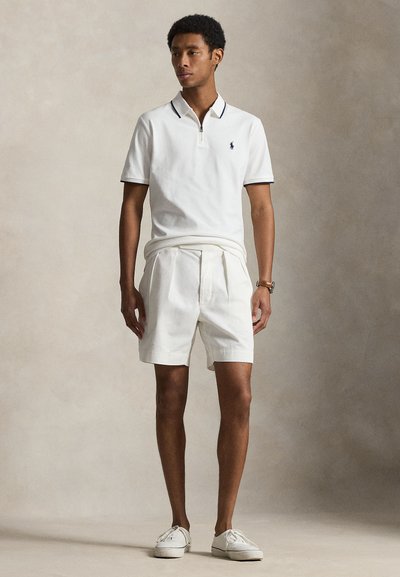 Jeune homme debout portant un polo blanc à manches courtes, un short blanc plissé, des baskets blanches et une montre-bracelet sur un fond neutre.