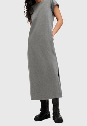Mujer que lleva un vestido midi gris sin mangas combinado con botas negras hasta la mitad de la pantorrilla con hebillas, de pie con las manos en los bolsillos del vestido.