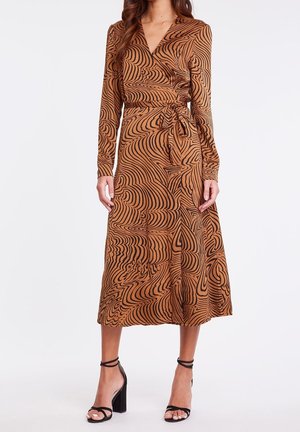 Femme portant une robe portefeuille marron à manches longues avec des motifs noirs tourbillonnants et des sandales à talons hauts noires, debout devant un fond blanc.