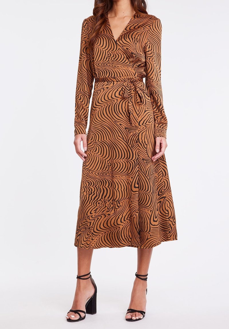 Femme portant une robe portefeuille marron à manches longues avec des motifs noirs tourbillonnants et des sandales à talons hauts noires, debout devant un fond blanc.