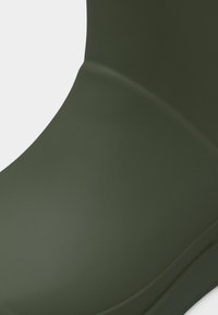 Κοντινό πλάνο μιας καμπυλωτής επιφάνειας σε ελαιί πράσινο με λεία υφή και ραφή, πιθανώς μέρος ενδύματος ή αξεσουάρ.