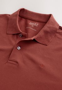 Camiseta tipo polo color óxido hecha de tela texturizada, con cuello, tapeta con dos botones y bordado de logotipo en el pecho.