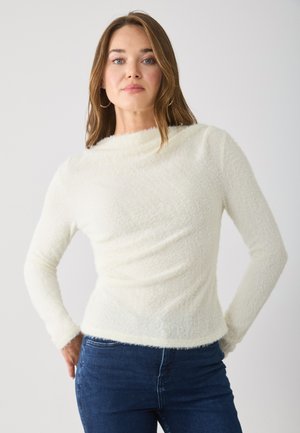 Hvit, teksturert strikkegenser med lange ermer og båtutringning. Stoffet ser mykt og lodden ut, kombinert med blå jeans.