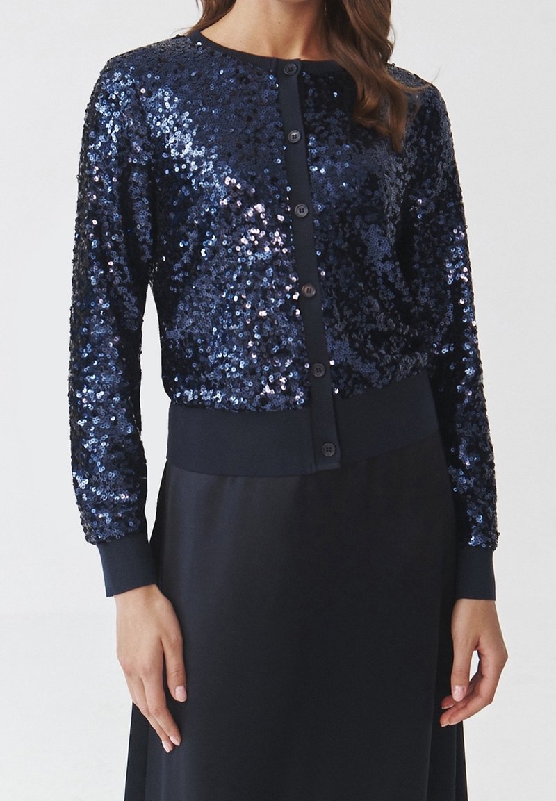 Femme portant un cardigan boutonné bleu marine à sequins avec des poignets et un ourlet côtelés, associé à une jupe foncée assortie.