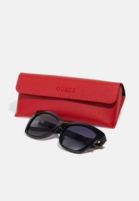 Black sunglasses with gradient lenses beside a red textured leather case featuring an embossed "GUESS" logo.