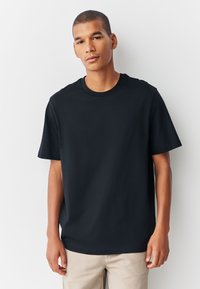 Juoda medvilninė marškinėliai su apvalia iškirpte, trumpomis rankovėmis, laisvo kirpimo ir lygios tekstūros, dėvimi su smėlio spalvos kelnėmis. Paprastas dizainas, be raštų.