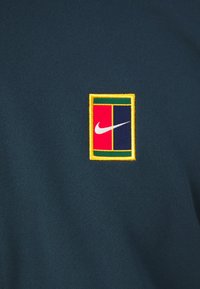 Sötétzöld póló, amelyen hímezett téglalap alakú logó található, piros, kék és sárga kerettel, középen pedig egy fehér Nike logóval.
