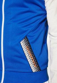 Blå zip-up jacka med vita accenter, i en texturerad tyg, sidoficka med ett svart och vitt mönster, samt orange detaljer.