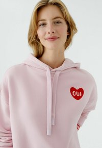 Sweat à capuche rose avec une poche avant, arborant un motif de cœur rouge et le mot « OUI » brodé en blanc. Tissu doux et cordon de serrage ajustable.