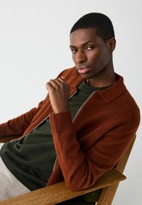 Rust-colored cardigan with ribbed cuffs over a green shirt, featuring a zip closure and a soft texture. Model seated in a wooden chair.