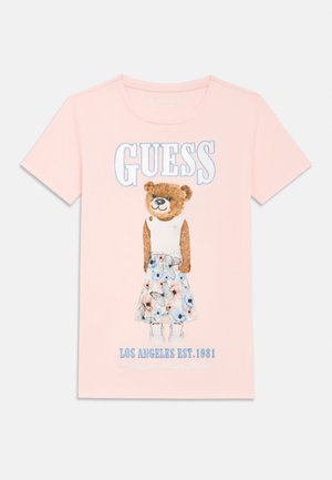 Hellrosa T-Shirt mit einem Teddy, der ein weißes Oberteil und einen geblümten Rock trägt, "GUESS"-Schriftzug darüber und "LOS ANGELES EST. 1981" darunter.