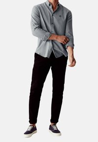 Camicia botton-down grigia chiara con maniche lunghe e logo ricamato scuro; abbinata a pantaloni neri e sneaker blu navy con suola bianca.