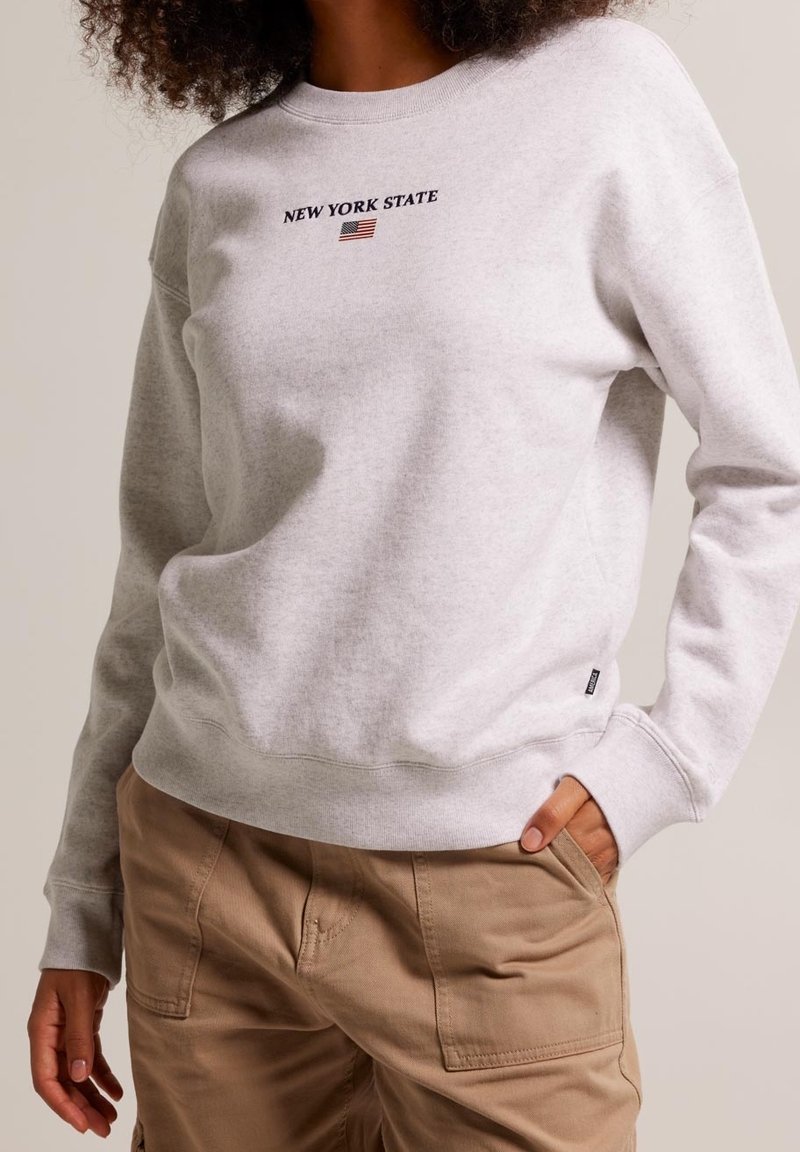 Sudadera gris claro con cuello redondo, que presenta el texto "ESTADO DE NUEVA YORK" y un gráfico de la bandera americana. Mezcla de algodón, textura suave, puños acanalados.