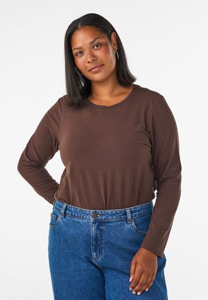 Femme à la peau brun moyen et aux cheveux noirs raides, portant une chemise marron à manches longues et un jean bleu, debout avec une main sur la hanche.