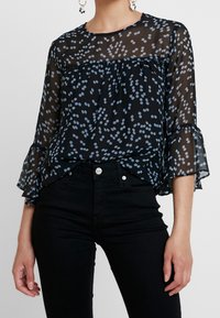 Schwarze durchsichtige Bluse mit hellblauen Punkten, rundem Ausschnitt und Rüschensleeves, kombiniert mit schwarzen Jeans.