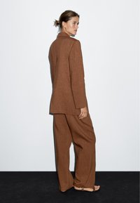 Brązowy lniany garnitur z długim żakietem i szerokimi spodniami; charakteryzuje się kołnierzykiem i luźnym krojem, prezentując teksturowany, naturalny materiał.
