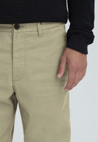 Beežid sirge lõikega püksid, ühtlase tekstuuriga ja ees taskuga, kombineeritud musta soonikkoega pikkade varrukatega särgiga.