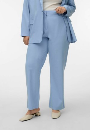 Kvinde iført lyseblå skræddersyede bukser og matchende blazer med cremefarvede mules med firkantet tå og lav hæl, stående mod en ensfarvet baggrund.