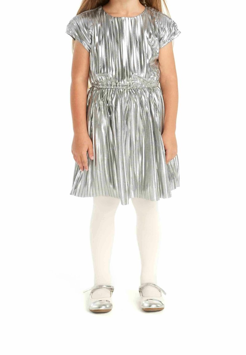 Enfant portant une robe plissée argentée, des collants blancs et des chaussures argentées, debout les mains détendues le long du corps.