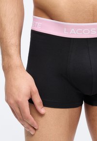 Άνδρας με κορμό που φοράει μαύρα μποξεράκια με ροζ ζωνάκι μέσης με την ετικέτα «Lacoste», το χέρι ακουμπάει στον μηρό πάνω σε απλό φόντο.