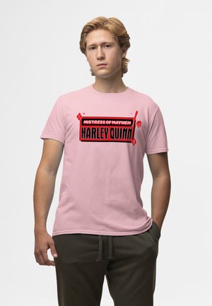 Giovane uomo con capelli chiari che indossa una maglietta rosa con la scritta "Mistress of Mayhem Harley Quinn" in rosso e nero, in piedi con una mano in tasca.
